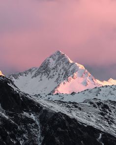 the mountains are covered in snow as the sun goes down on them and pink clouds loom over them