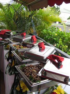 many trays of food are lined up on a table with red napkins and green plants