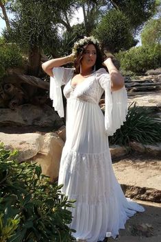 a woman wearing a white dress and holding her hands on her head while standing in front of some trees