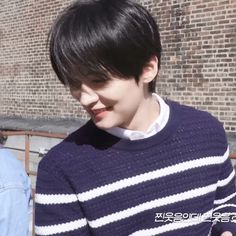 a young man with black hair wearing a blue and white striped sweater standing in front of a brick wall