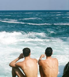 two men sitting on the rocks looking out at the ocean