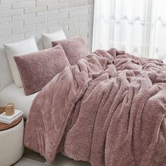 a bed with pink comforter and pillows in a white room next to a window