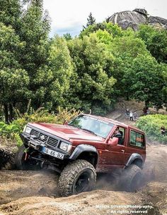 a red truck driving down a dirt road next to trees and people in the background