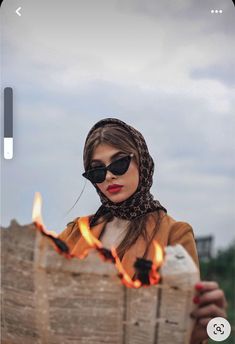 a woman wearing sunglasses and scarf holding a newspaper with flames in front of her face