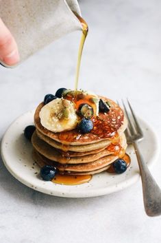 a person pouring syrup on pancakes with blueberries and bananas