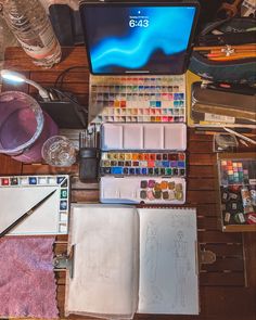 an open laptop computer sitting on top of a wooden desk covered in art supplies and paint