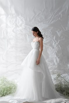 a woman in a white wedding dress standing against a backdrop with green plants and grass