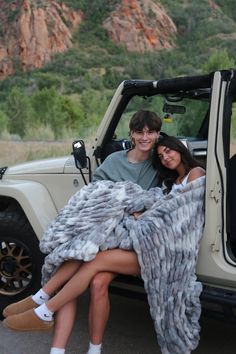 two people sitting in the back of a jeep with a blanket over their shoulders and smiling