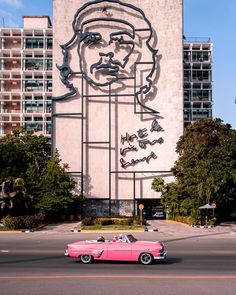 a pink car driving past a tall building with a giant poster on it's side