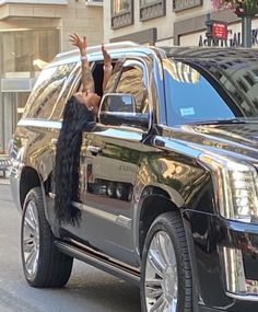 a woman is waving out the window of a black suv in front of a building