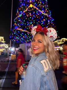 a woman in denim jacket standing next to a christmas tree with mickey mouse ears on it