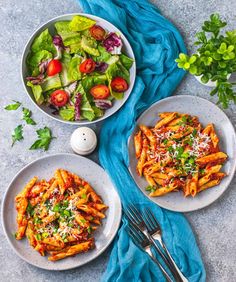 two plates of pasta and salad on a blue table cloth next to a bowl of salad