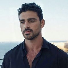 a man standing in front of the ocean wearing a black shirt and looking at the camera