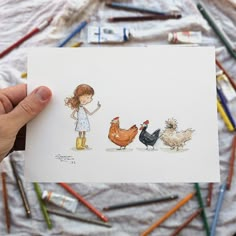 a person holding up a card with two chickens and one chicken on it in front of colored pencils