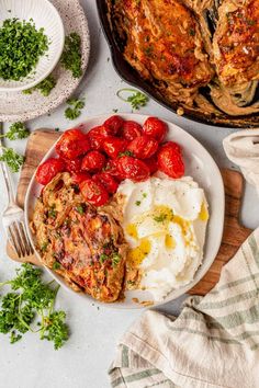 a white plate topped with meat, mashed potatoes and tomatoes next to a skillet