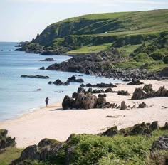 a person standing on the beach next to some rocks and water with green hills in the background