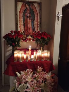 candles and flowers on a table in front of a virgin mary painting with red cloth