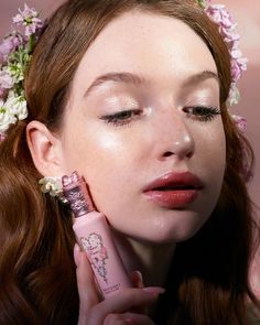 a woman with flowers in her hair is holding a tube of lipstick to her face