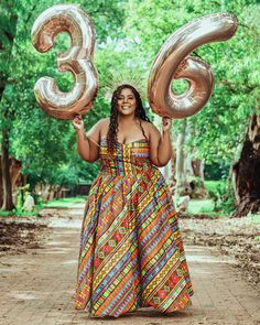 a woman in a colorful dress holding two large gold balloons and smiling at the camera