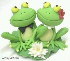 two green frog figurines sitting on top of each other with flowers in their mouths