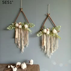 two wall hangings with cotton flowers and greenery on them, next to a wooden table