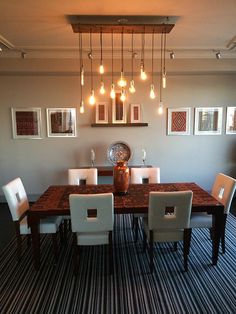 a dining room table with white chairs and pictures on the wall above it, along with hanging lights