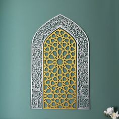 an intricately designed wall hanging on the side of a green wall next to a vase with flowers