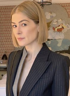 a woman with blonde hair wearing a pinstripe suit and grey shirt standing in front of a bed