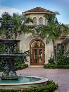 a fountain in front of a house with palm trees and bushes around it on a brick walkway