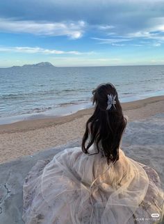 a woman sitting on top of a sandy beach next to the ocean wearing a white dress