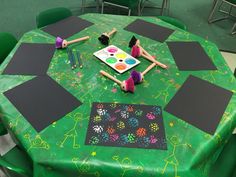 two dolls sitting at a green table with black paper on it and some paintbrushes