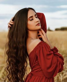 a woman with long hair in a red dress standing in a field looking off to the side