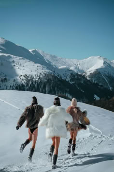 three women in fur coats are walking through the snow with mountains in the back ground