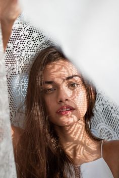 a woman with long brown hair standing in front of a white lace covered curtain and looking at the camera