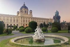 a fountain in front of a large building with statues on it's sides and trees around the perimeter