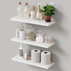 three white shelves holding toilet paper, vases and other bathroom accessories on top of each shelf