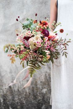 a woman in white dress holding a bouquet of flowers and greenery against a stone wall