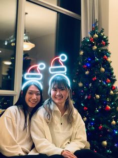 two women sitting next to each other in front of a christmas tree with lights on it