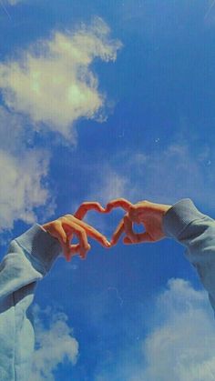 two hands making a heart shape with their fingers against a blue sky and white clouds