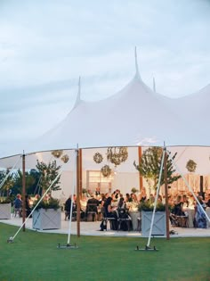 a group of people sitting at tables under a tent with white walls and poles in the grass