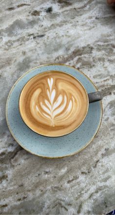 a cappuccino on a saucer sitting on a marble counter top next to a person's hand