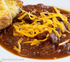 a white plate topped with chili and cheese next to a piece of bread on top of it