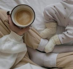 a woman holding a cup of coffee while laying in bed with her feet on the pillow