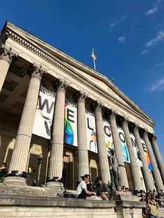 people are sitting on the steps in front of a building with columns and flags hanging from it