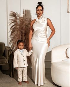 a woman standing next to a little boy in a white dress and suit with one hand on his hip