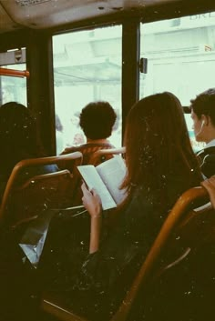 two people are sitting on a bus and one is reading a book while the other looks out the window