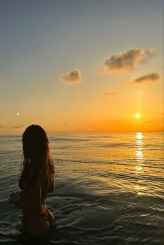 a woman sitting in the water at sunset with her back turned to the camera,