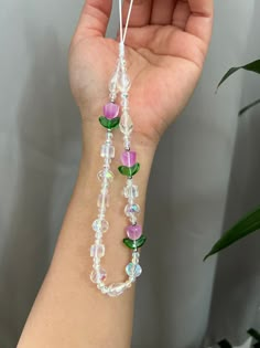 a hand holding a beaded bracelet with flowers and leaves attached to it, in front of a plant