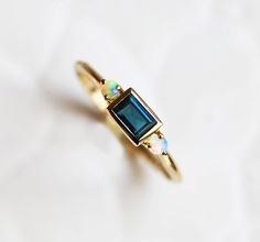 Modern and simple baguette teal sapphire ring with an australian opal on each side. Beautiful as an engagement or wedding ring. Available in 14k and 18k rose/white/yellow gold, platinum. Please select the metal and ring size from the drop-down menu. If you would like to have other gemstones instead of diamonds/opals please contact us before purchase regarding pricing. Item details: teal baguette sapphire - 4x 2.5mm approx Australian opal on each side 14k/18k solid gold, platinum The ring will be Teal Engagement Ring, Round Blue Sapphire Ring, Teal Sapphire Ring, Engagement Ring Three Stone, Twig Engagement Ring, Ring Three Stone, Blue Opal Ring, Multi Gemstone Ring, Teal Sapphire