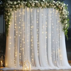 a white curtain with lights and flowers on the top is decorated for a wedding ceremony